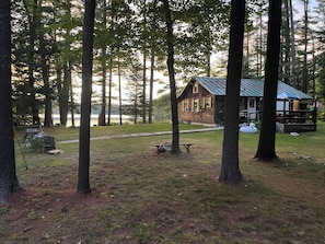A cabin with a deck, surrounded by trees and a grassy area with a picnic table. - Charming 3-bedroom cabin on David Pond (Fayette)