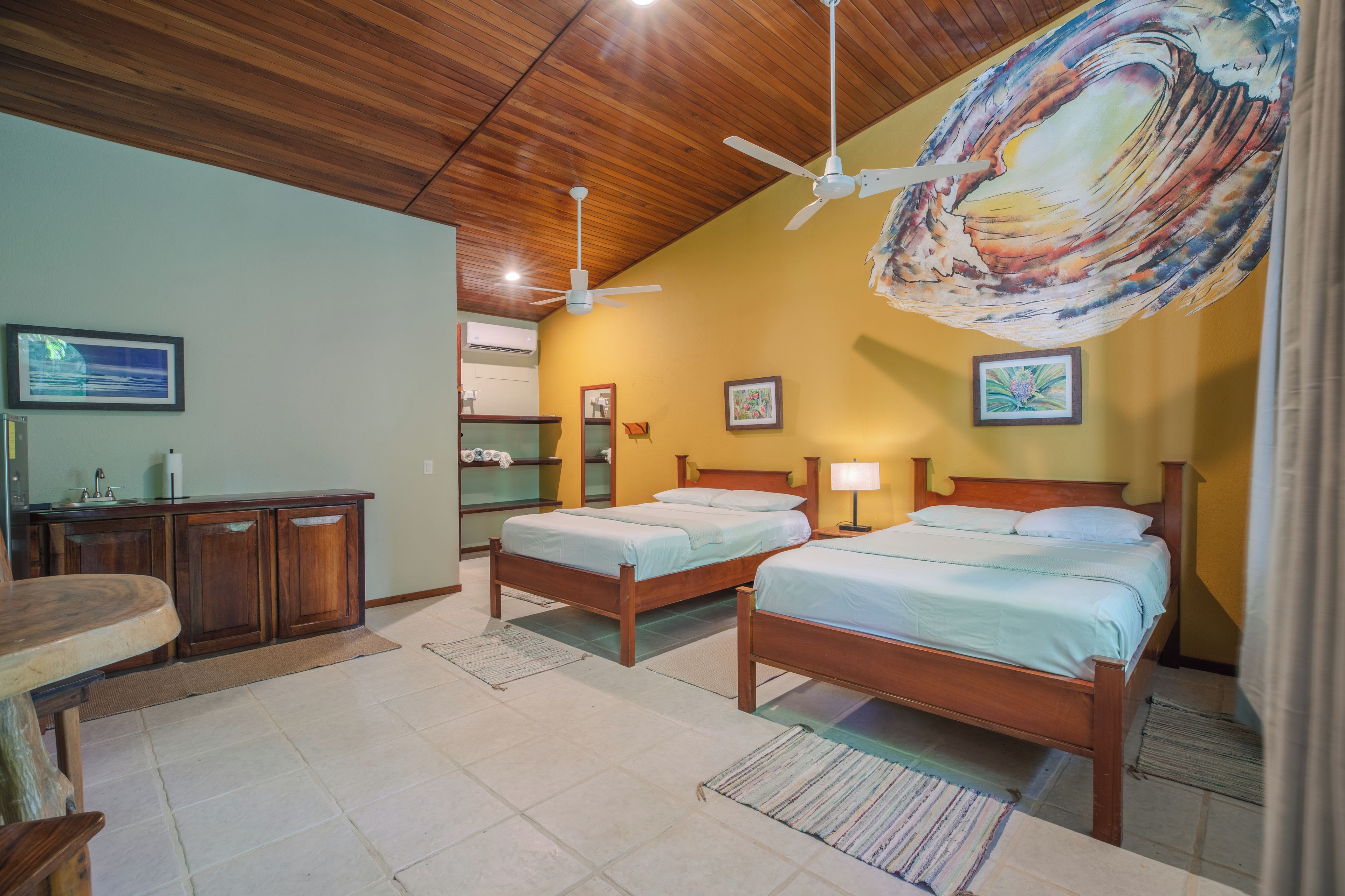 Habitación de hotel con dos camas de madera, un escritorio de madera, un ventilador de techo y un cuadro abstracto grande en la pared.