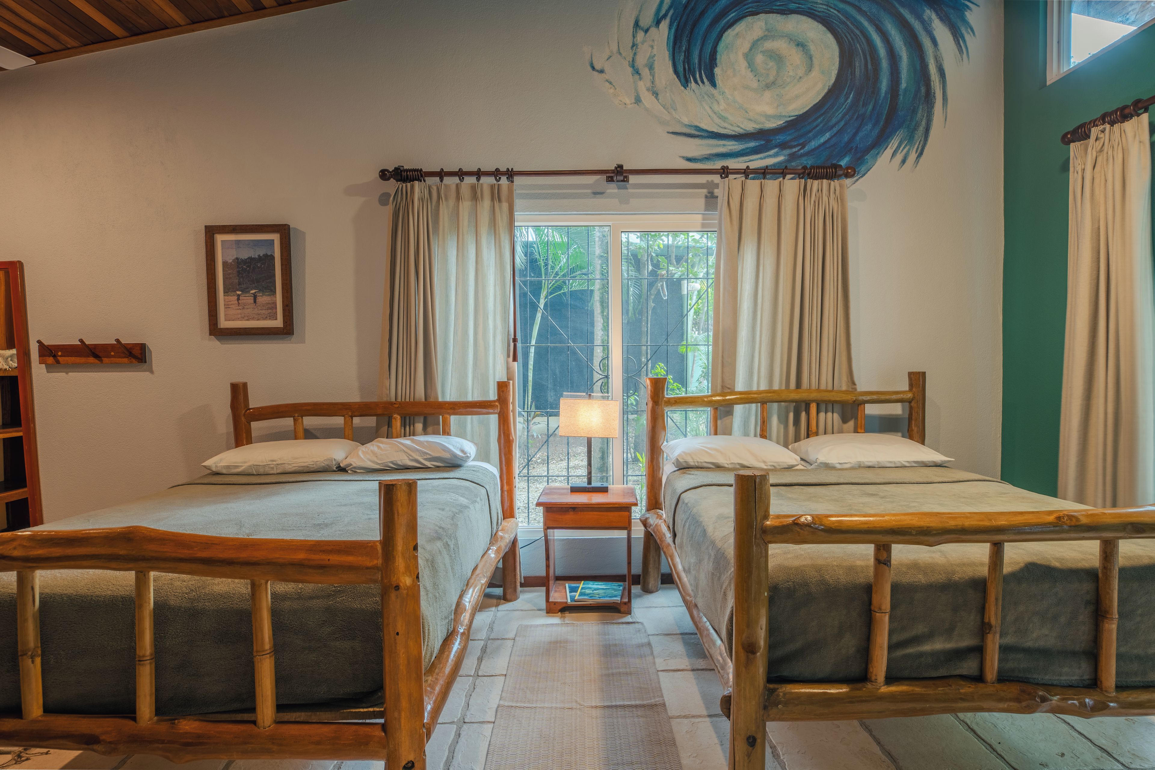 Una habitación con dos camas de madera, una mesita de noche pequeña con lámpara, un cuadro en la pared y una ventana con cortinas.