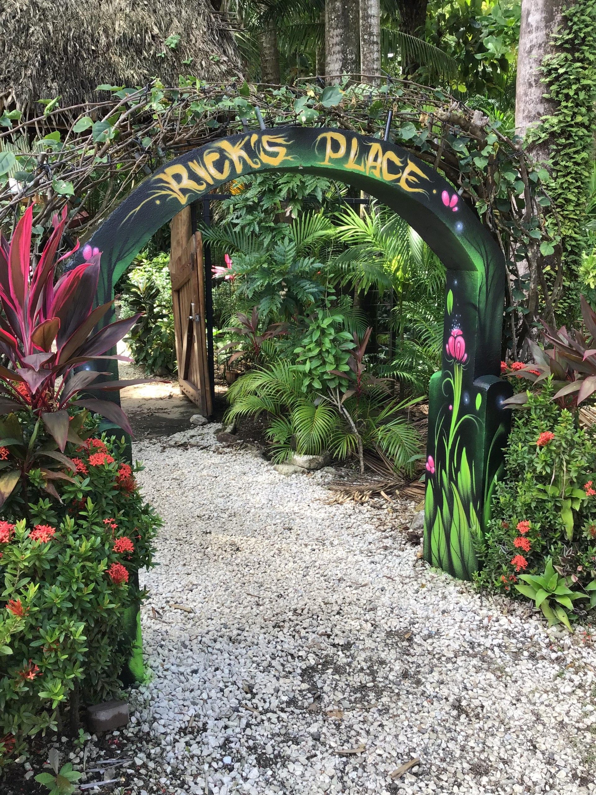 An entrance archway with the inscription 'Rick's Place' surrounded by lush greenery and flowering plants.