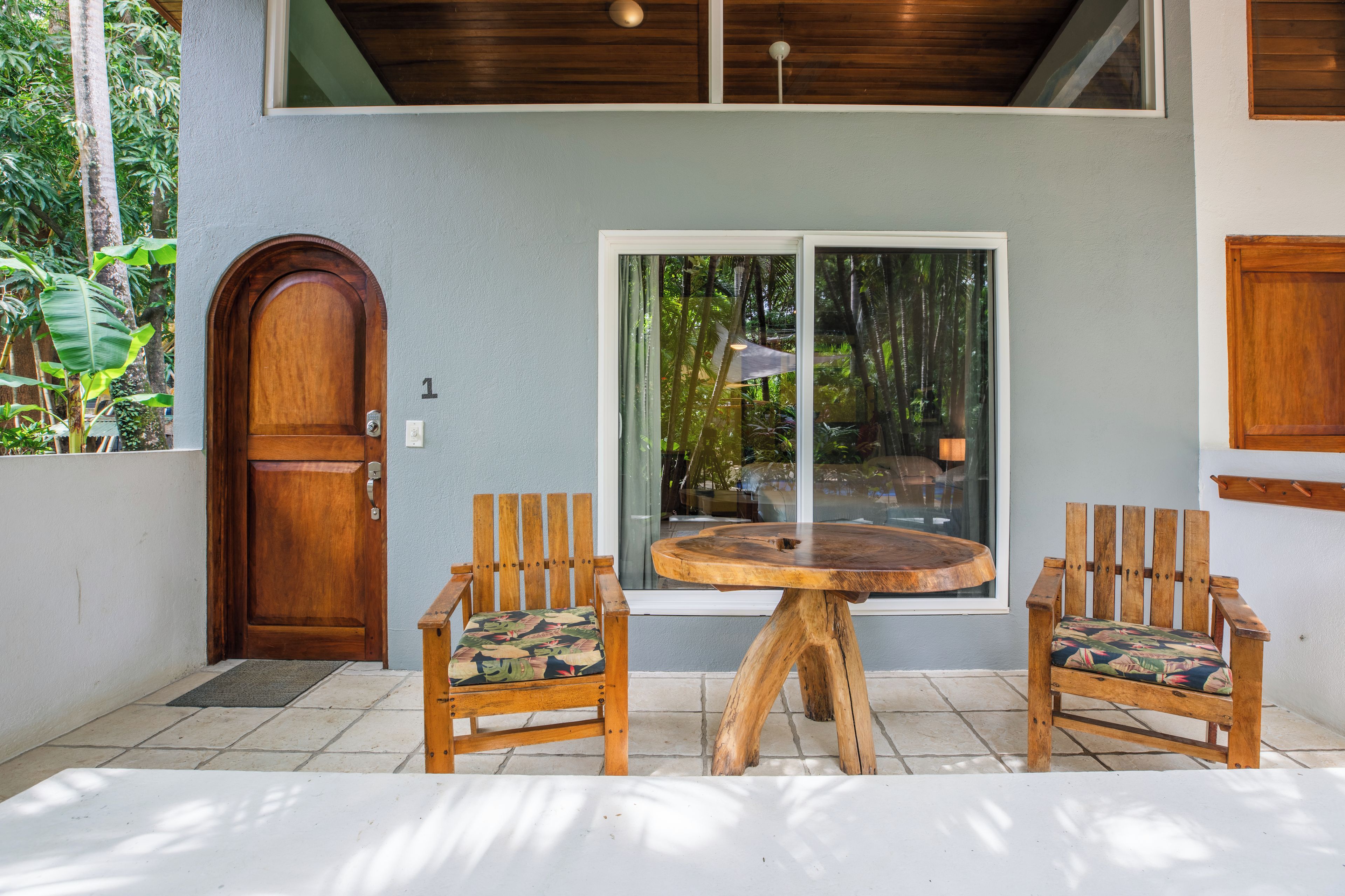 A wooden door, a window with curtains, and two wooden chairs with a round table in a covered outdoor area.