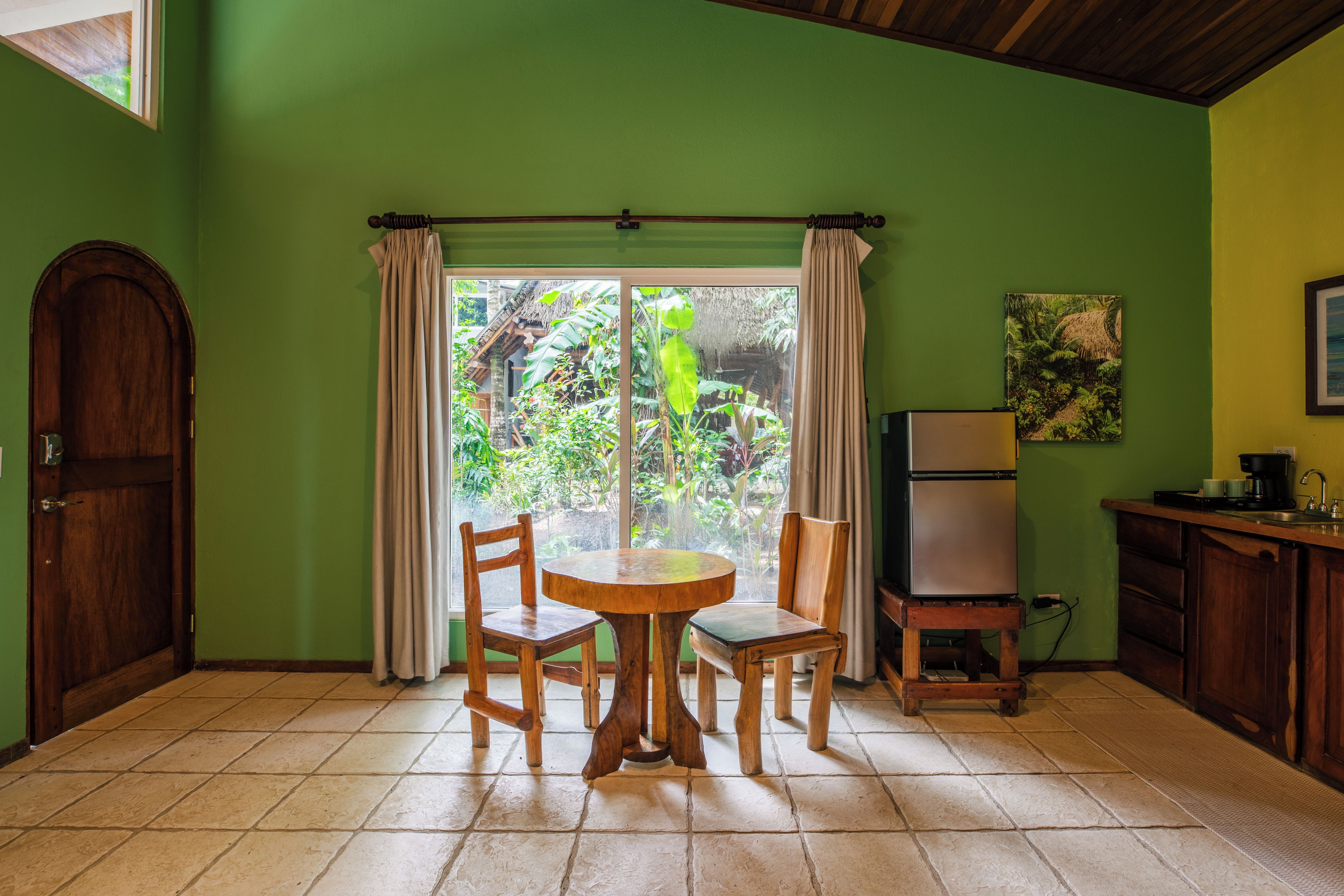 Una habitación con puerta de madera, una mesa con sillas, un refrigerador y una ventana con cortinas.