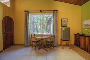 A room with a wooden door, a window with curtains, a round table with chairs, a refrigerator, and a painting on the wall. - Rick's Place (Nosara)