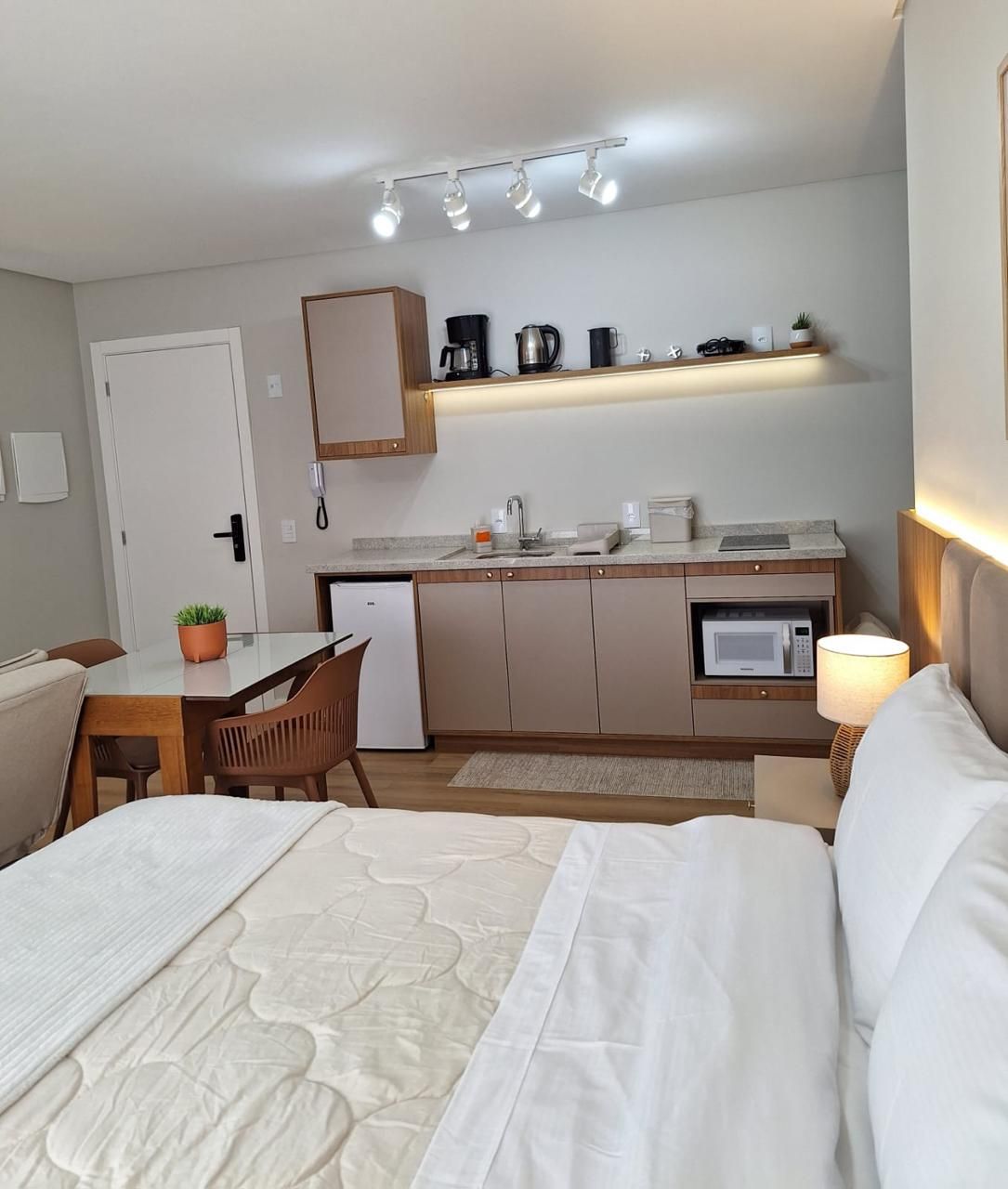 Una habitación de hotel con cama, escritorio con silla, kitchenette con microondas, cafetera y estante con varios objetos.