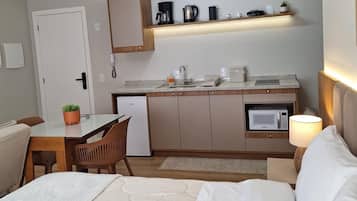 Una habitación de hotel con cama, escritorio con silla, kitchenette con microondas, cafetera y estante con varios objetos.