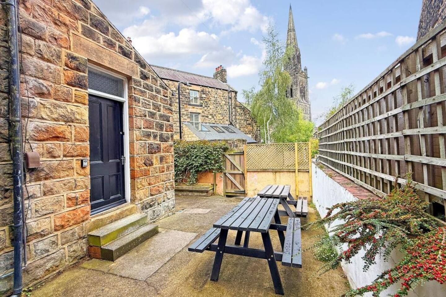 A stone building with a black door, a wooden fence, and a picnic table in a courtyard.