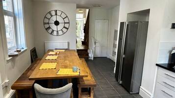 A kitchen with a wooden table, chairs, a large wall clock, and a refrigerator.