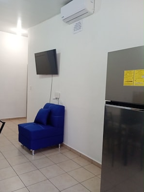A room with a blue sofa, a flat-screen TV, a refrigerator, and an air conditioning unit.