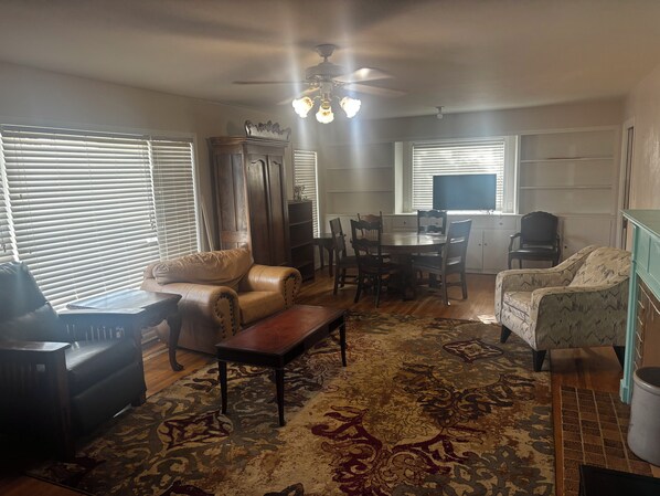 A living room with a sofa, a coffee table, a patterned rug, a dining table with chairs, and a ceiling fan.