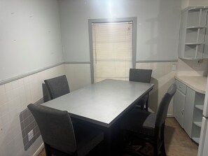 A small, enclosed dining area with a table and four chairs, a window with blinds, and a kitchenette with cabinets.