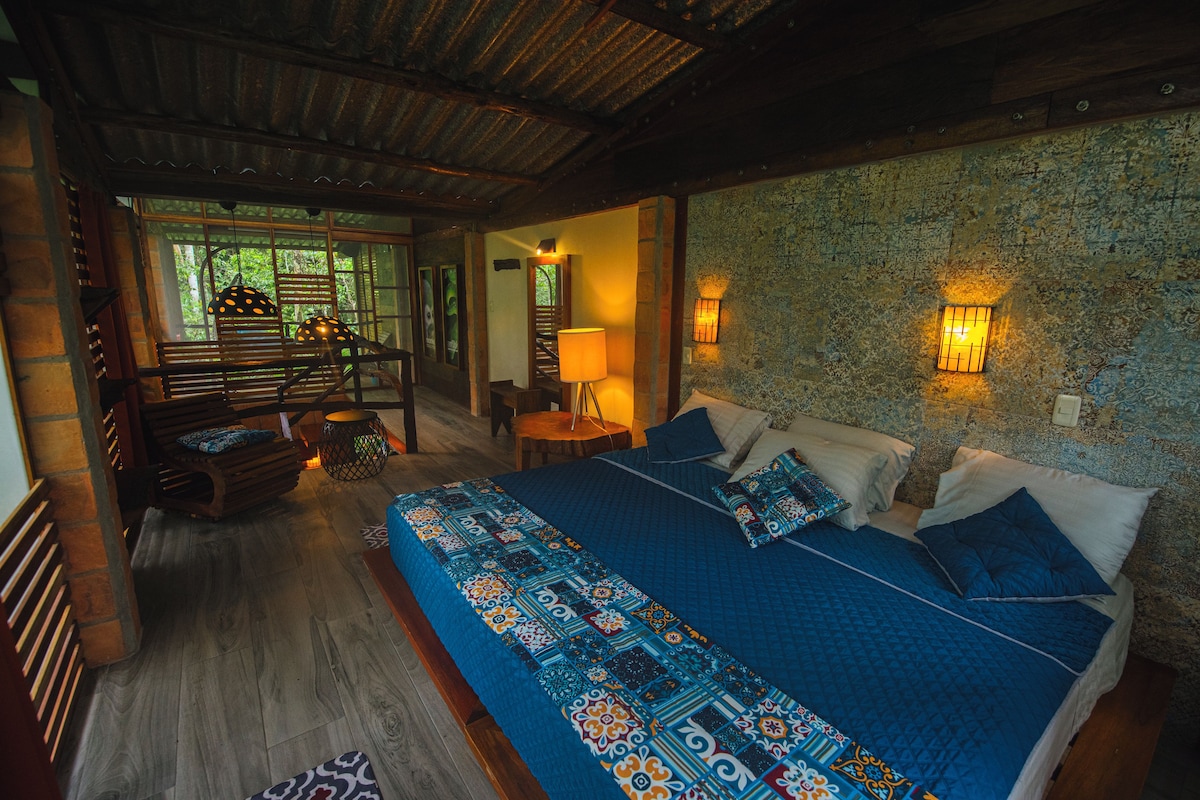 A bedroom with a large bed, a wooden bench, a small table, and a wall-mounted lamp.