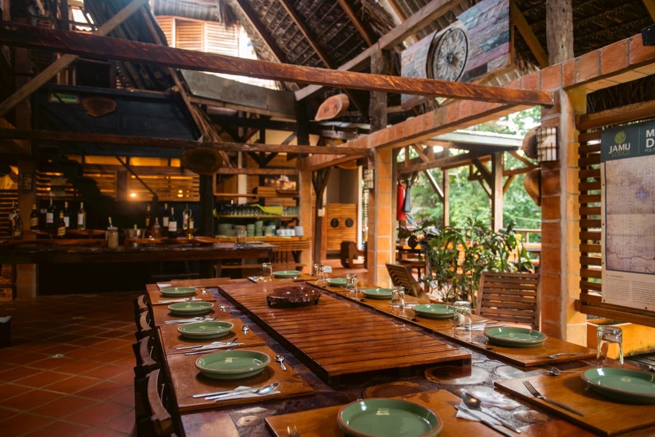 Ein Restaurant mit langen Holztischen, gedeckt für das Essen, einem Barbereich mit Flaschen und Blick auf Grünflächen draußen.
