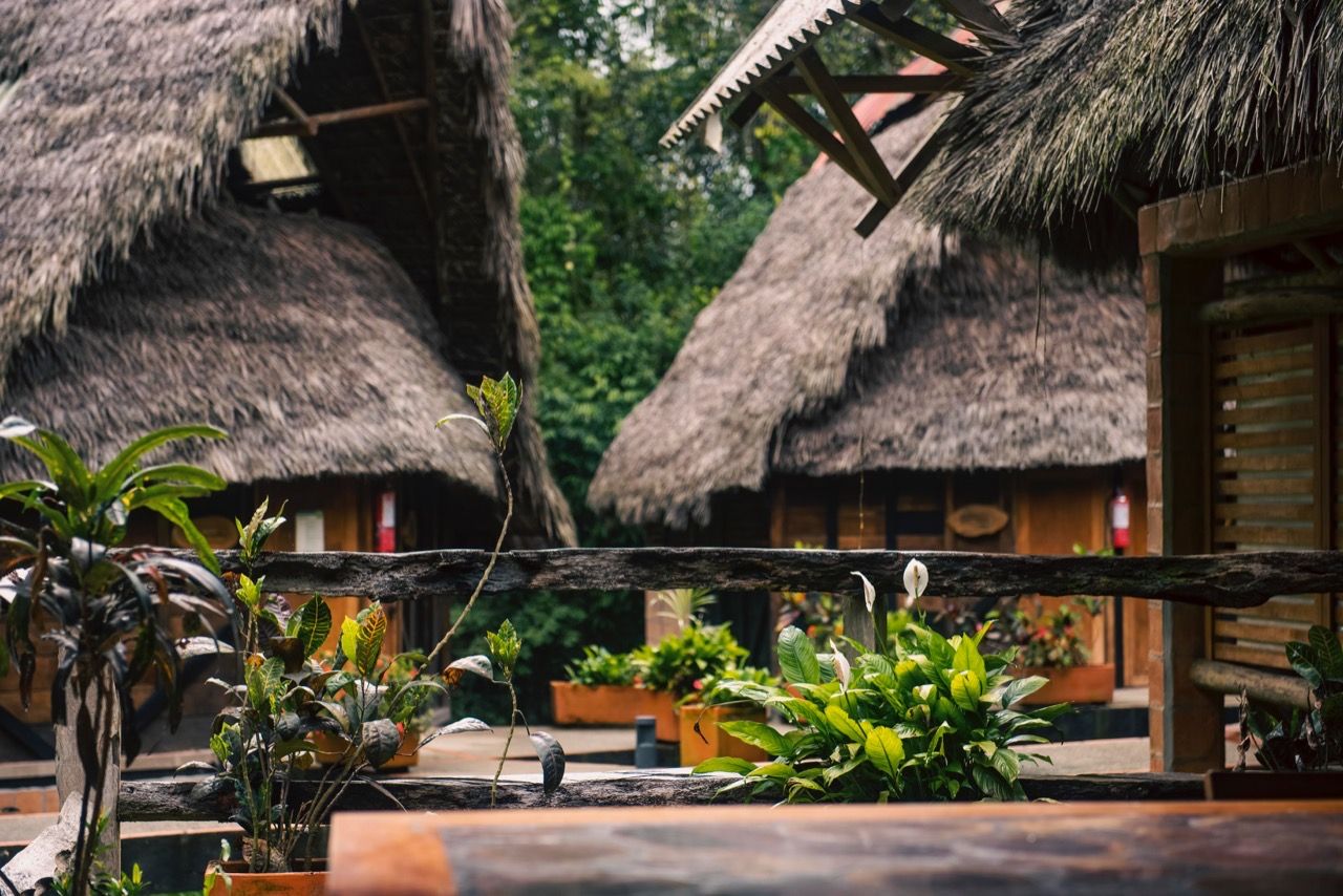 Cabanas com telhado de palha, balaústres de madeira e plantas em vasos no primeiro plano.