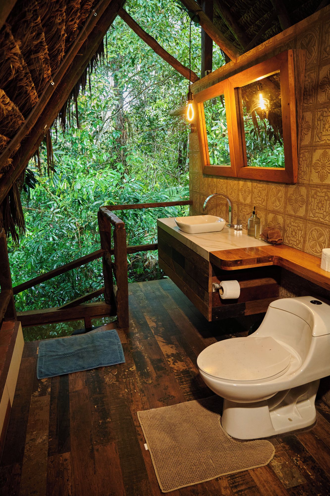 Ein rustikales Badezimmer mit Holzboden, Toilette in Weiß, Waschbecken mit Spiegel und Fenster mit Blick auf dichtes Grün.