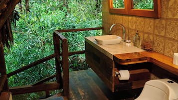 Ein rustikales Badezimmer mit Holzboden, Toilette in Weiß, Waschbecken mit Spiegel und Fenster mit Blick auf dichtes Grün.