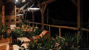 Un área de estar al aire libre con mobiliario de madera y plantas en macetas, bajo una estructura de madera con vigas a la vista y luces colgantes.