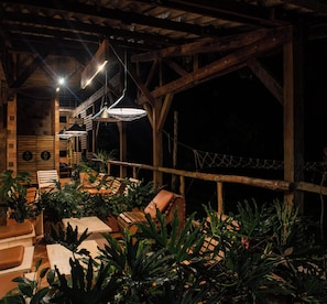 Uma área de estar ao ar livre com móveis de madeira e plantas em vasos, sob uma estrutura de madeira com vigas aparentes e luminárias pendentes.