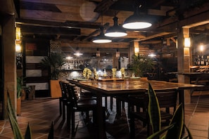 A restaurant with a long wooden table, multiple chairs, hanging pendant lights, and a tiled floor.
