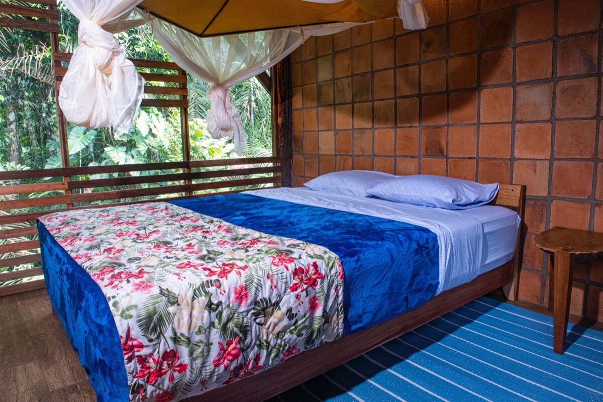 A room with a bed, a window with a view of greenery, and a wooden floor.