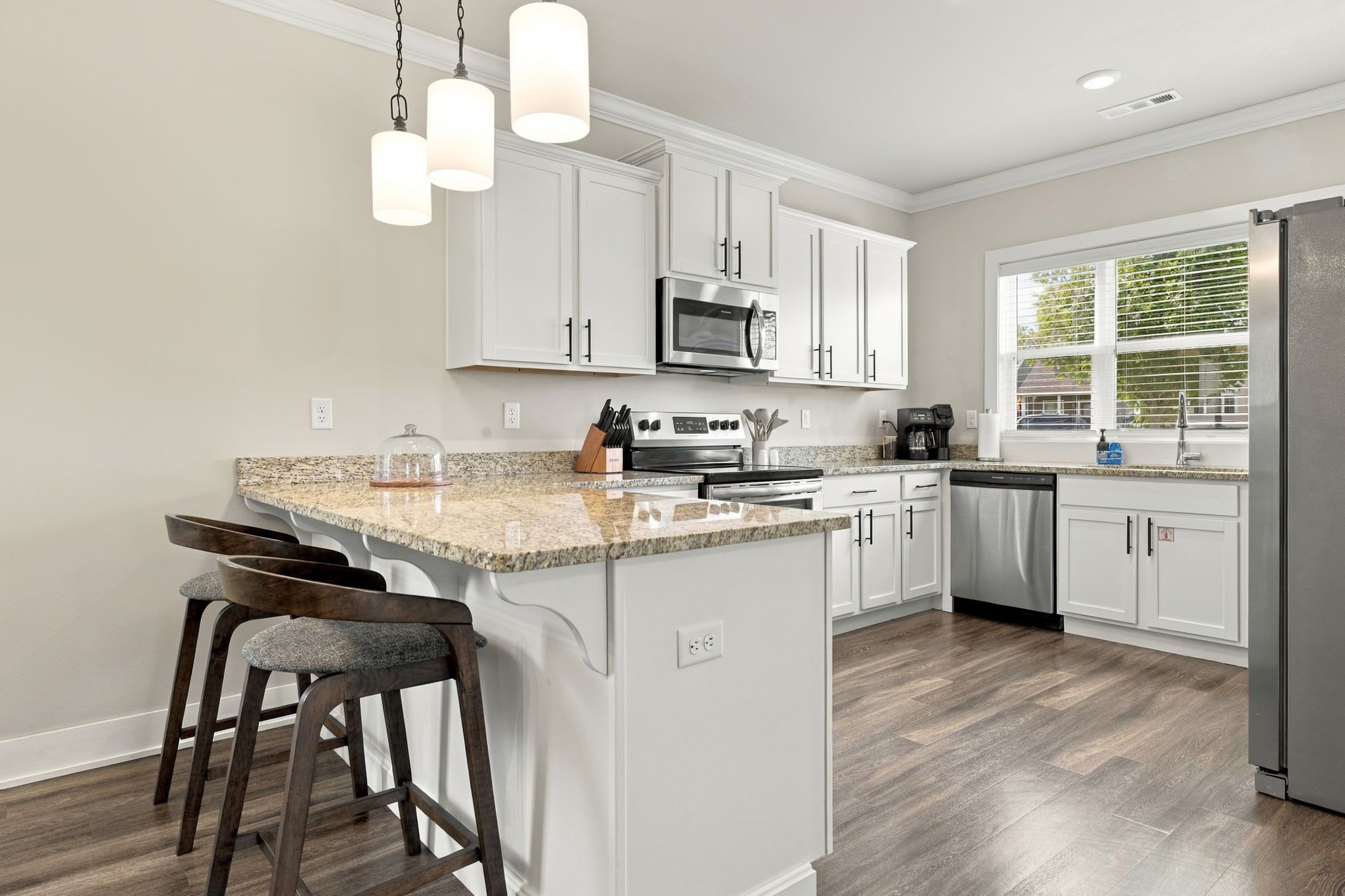 A modern kitchen with white cabinets, a granite countertop, a stainless steel refrigerator, and a window with blinds.
