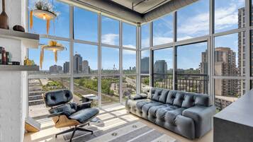A modern living room with a sofa and a chair, large windows with a city view, and a hanging light fixture.