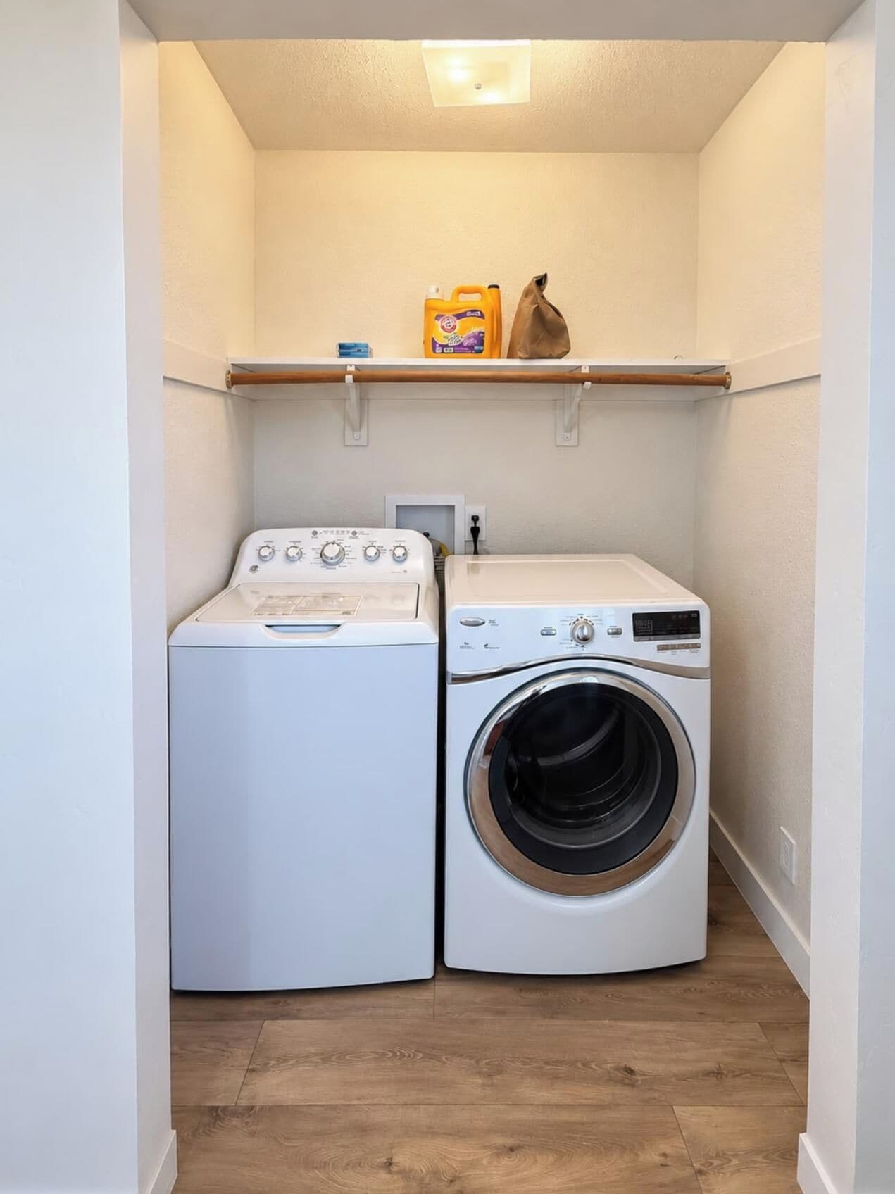 A laundry room with a washing machine and dryer, wooden flooring, and a shelf with laundry supplies.