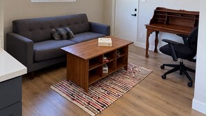 A living room with a sofa, coffee table, and a desk with a chair. There is a window with a view of greenery outside.