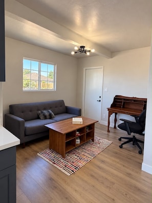Un salon comprenant un canapé, une table basse et un bureau avec une chaise. Une fenêtre offre une vue sur la verdure à l’extérieur.