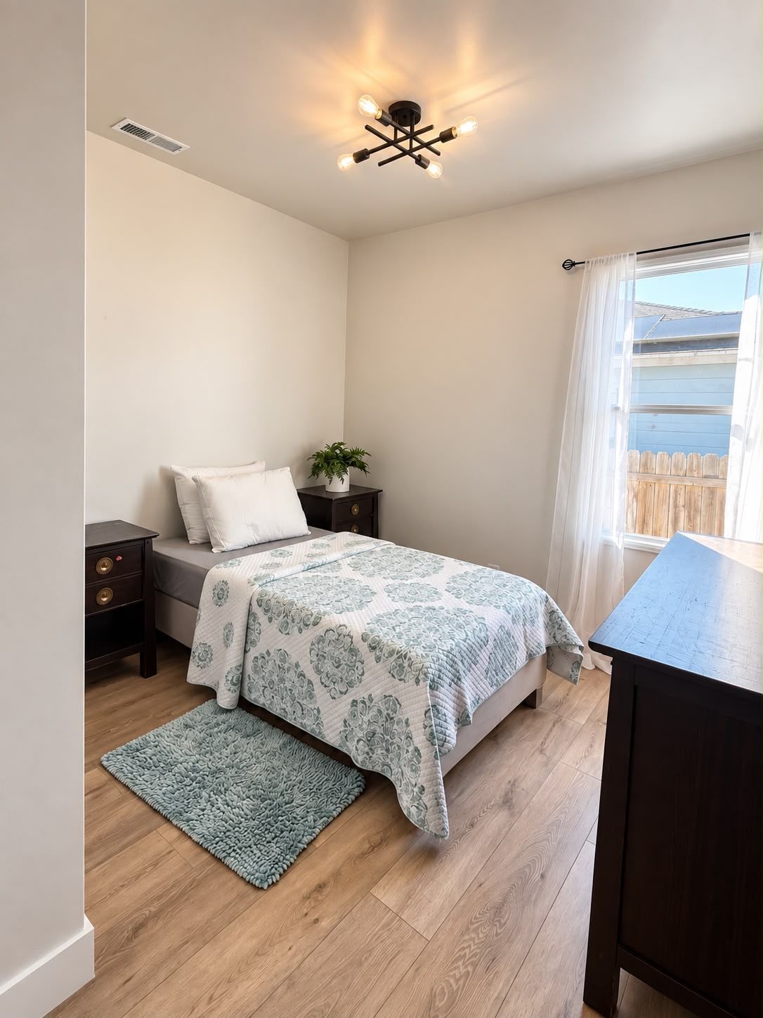 A bedroom with a bed, bedside tables, a nightstand, a window with curtains, a ceiling light fixture, and a small potted plant.