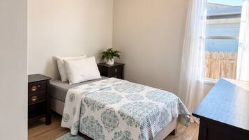 A bedroom with a bed, bedside tables, a nightstand, a window with curtains, a ceiling light fixture, and a small potted plant.