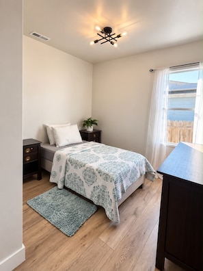 A bedroom with a bed, bedside tables, a nightstand, a window with curtains, a ceiling light fixture, and a small potted plant.