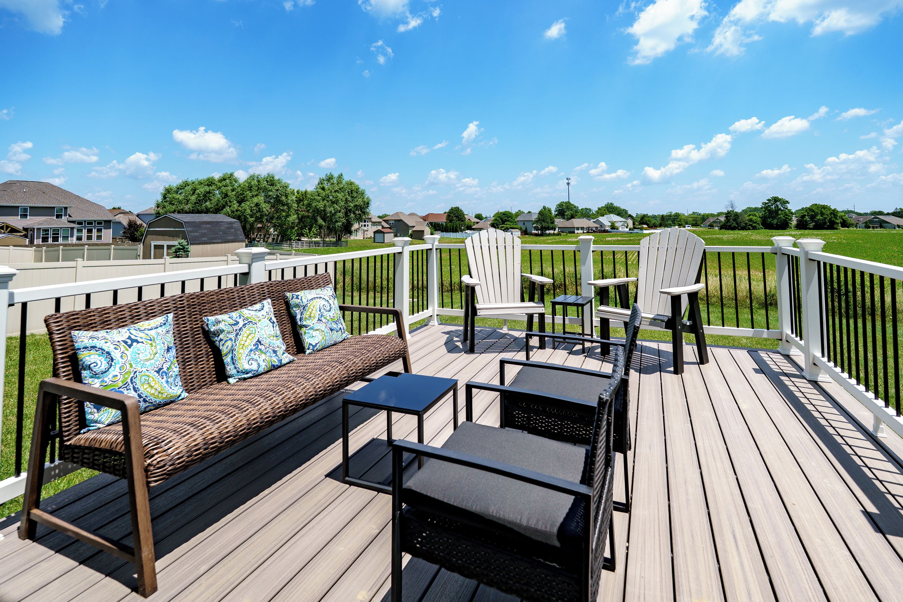 A wooden deck with outdoor furniture, including a sofa and chairs, overlooking a grassy area with houses in the background.