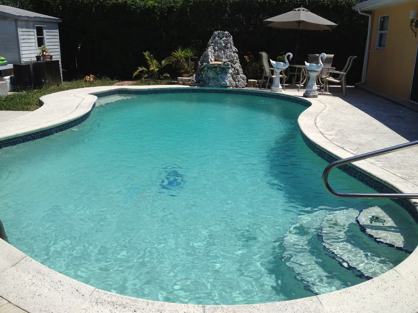 A clear swimming pool with a stone feature in the center, surrounded by a patio with chairs and an umbrella.