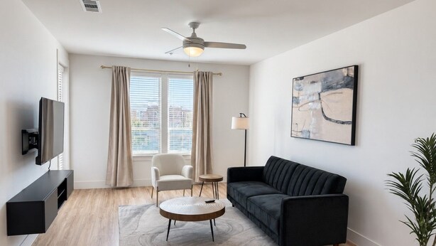 A modern living room with a flat-screen TV, a dark sofa, a round coffee table, a white armchair, a floor lamp, and a painting on the wall. - Modern 1BR Apartment near PGA and Universal Studios (Frisco)