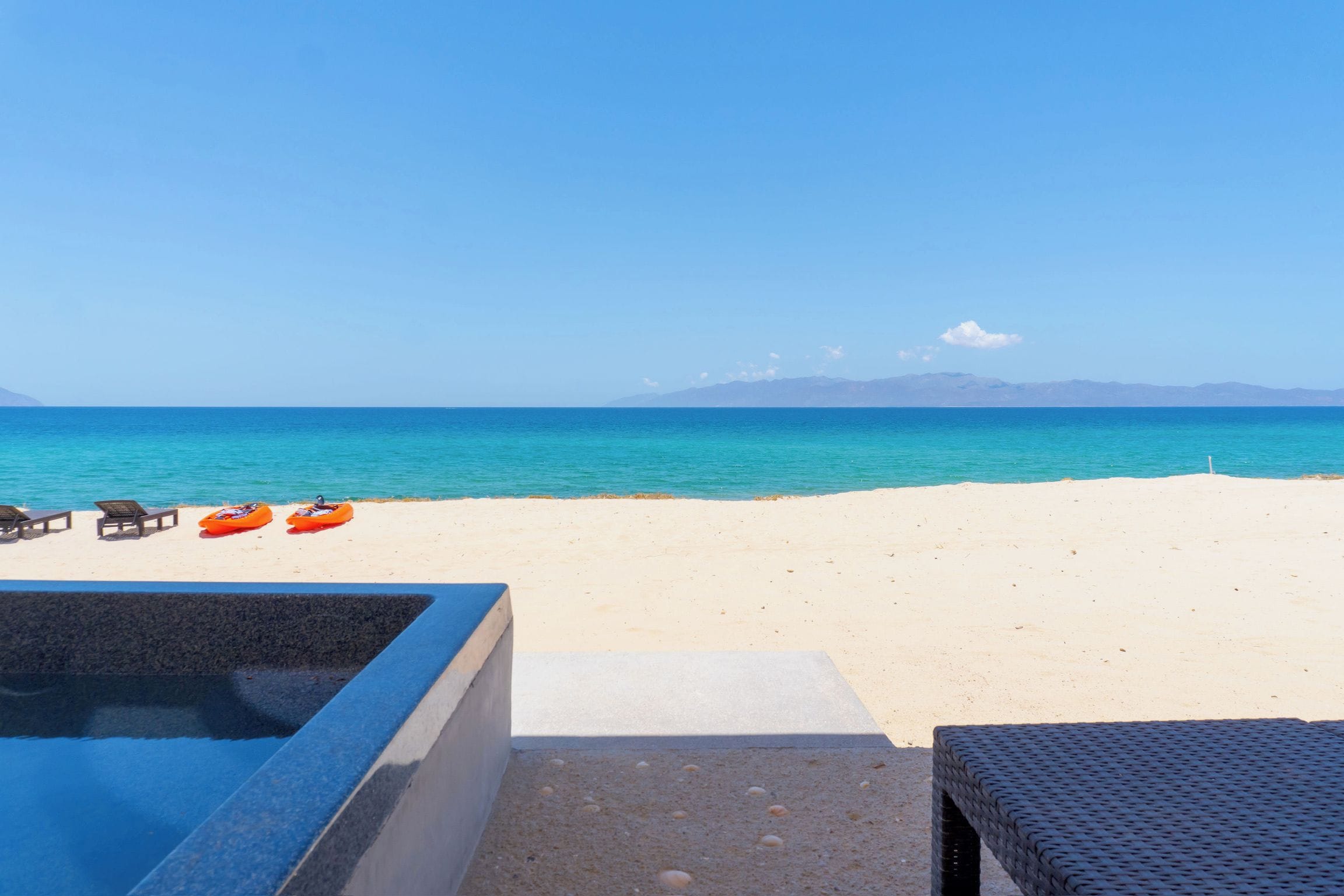 A beach with a clear view of the ocean and distant mountains. There are lounge chairs and an inflatable raft on the sand.