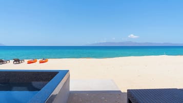 A beach with a clear view of the ocean and distant mountains. There are lounge chairs and an inflatable raft on the sand.