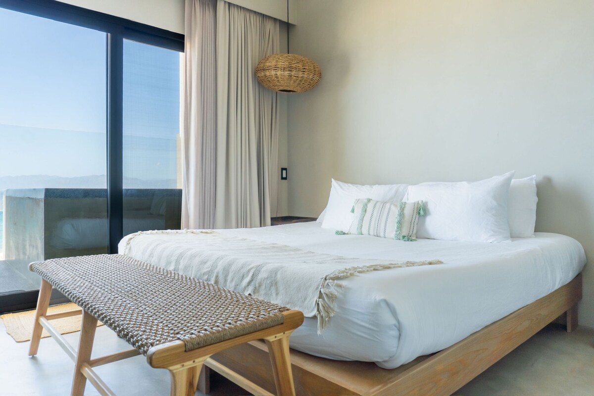 A hotel room with a large bed, a bench, a woven light fixture, and a view through a large window.