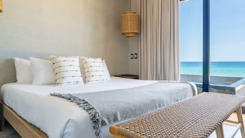 Un dormitorio con una cama grande, una mesita de noche, una silla de mimbre, vista al mar y una lámpara colgante.