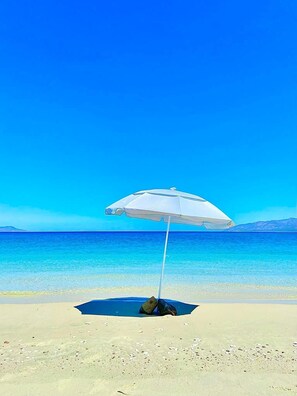 Escena de playa con cielo azul despejado, orilla arenosa y una sombrilla blanca que da sombra.