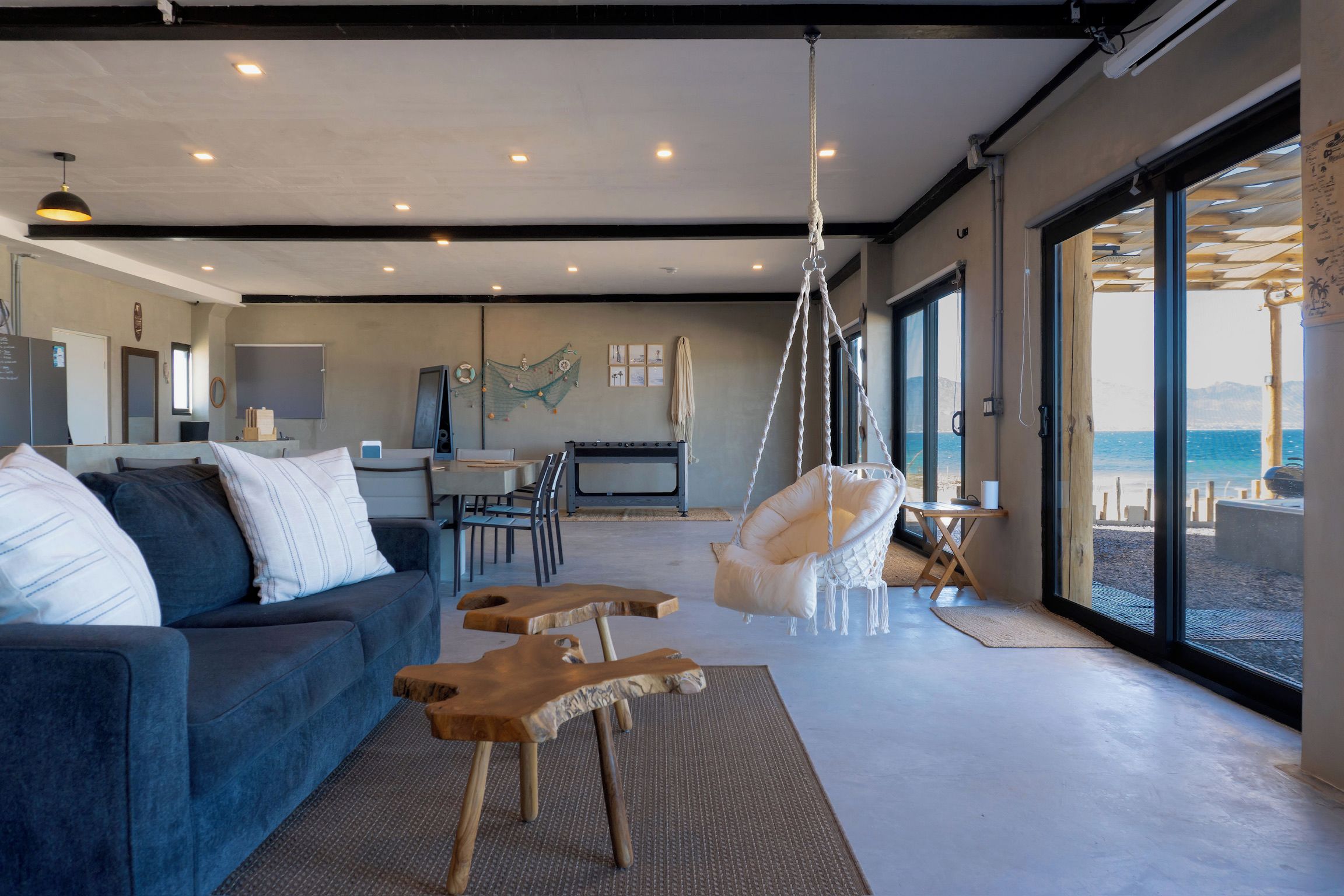 A modern living room with a sofa, chairs, a hanging chair, and a view of the sea through large windows.