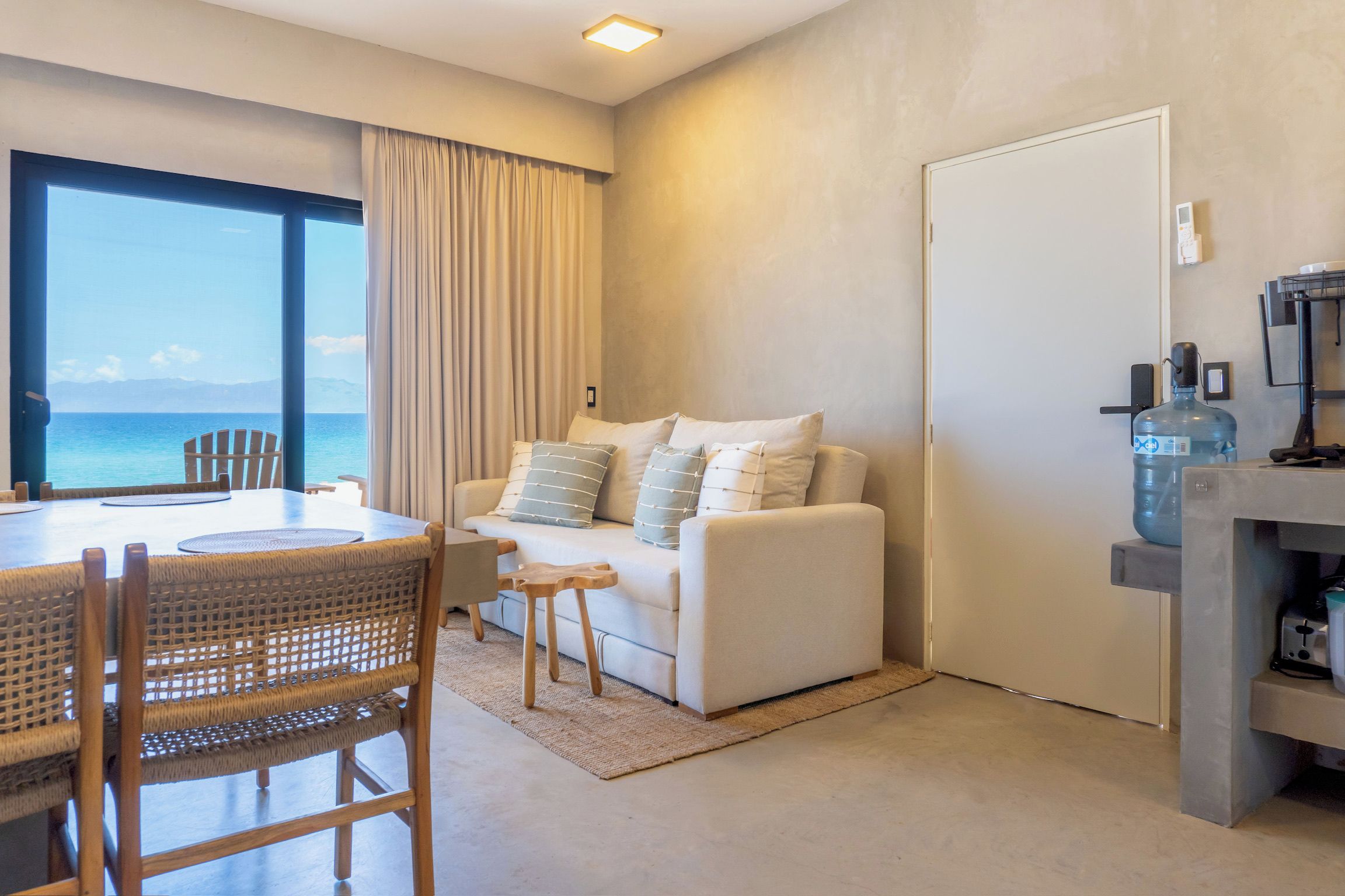 A modern living room with a white sofa, a dining table with chairs, a large window with a view of the sea, and a water dispenser on the right.