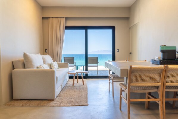 Una sala de estar moderna con un sofá, una mesa de comedor con sillas y vistas al mar a través de un ventanal grande.