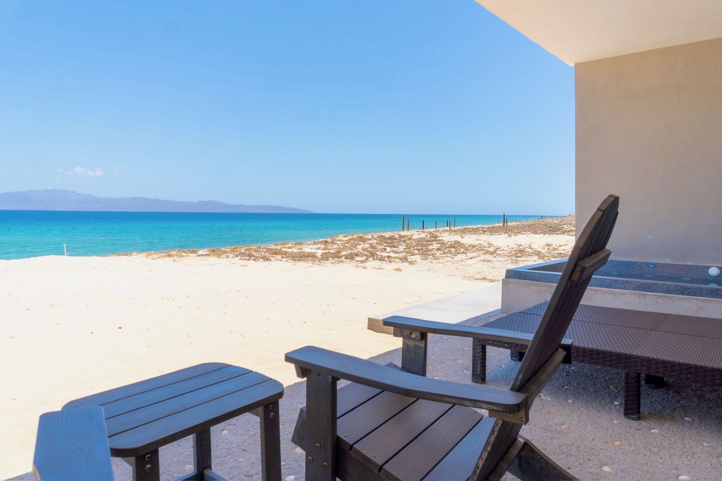 Vista a la playa con una silla y una mesa de madera, cielo despejado y mar tranquilo.