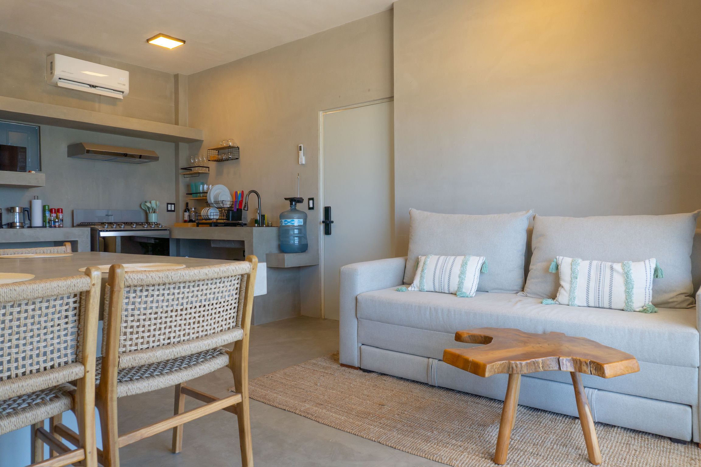 A modern living room with a sofa, a dining table, and chairs. There is a kitchen area with a stove, a water dispenser, and a microwave.