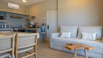 A modern living room with a sofa, a dining table, and chairs. There is a kitchen area with a stove, a water dispenser, and a microwave.