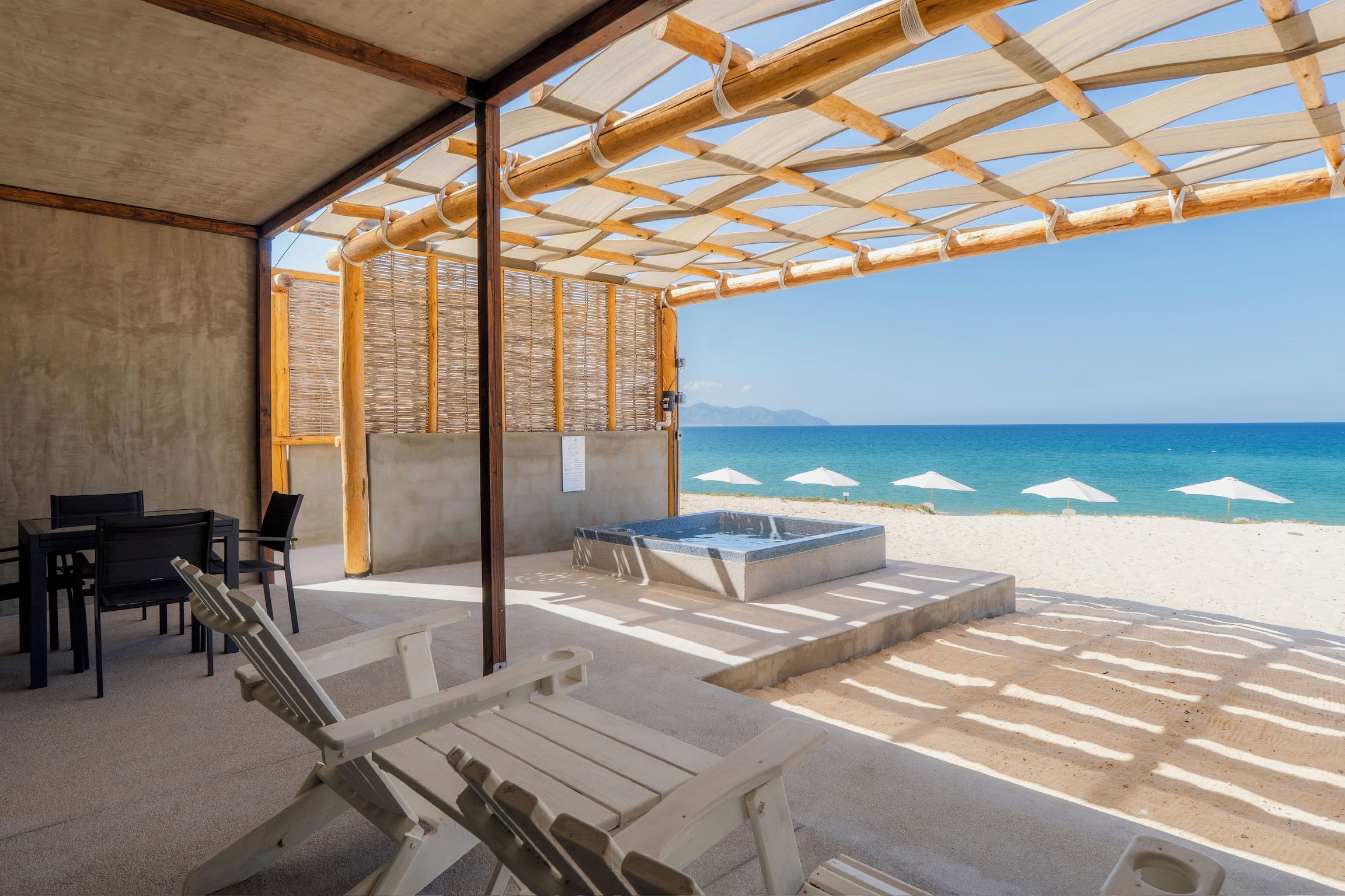 An outdoor seating area with lounge chairs and a hot tub, overlooking a beach with umbrellas.