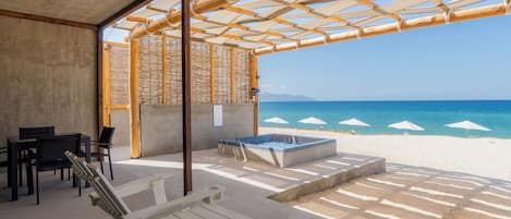 An outdoor seating area with lounge chairs and a hot tub, overlooking a beach with umbrellas.