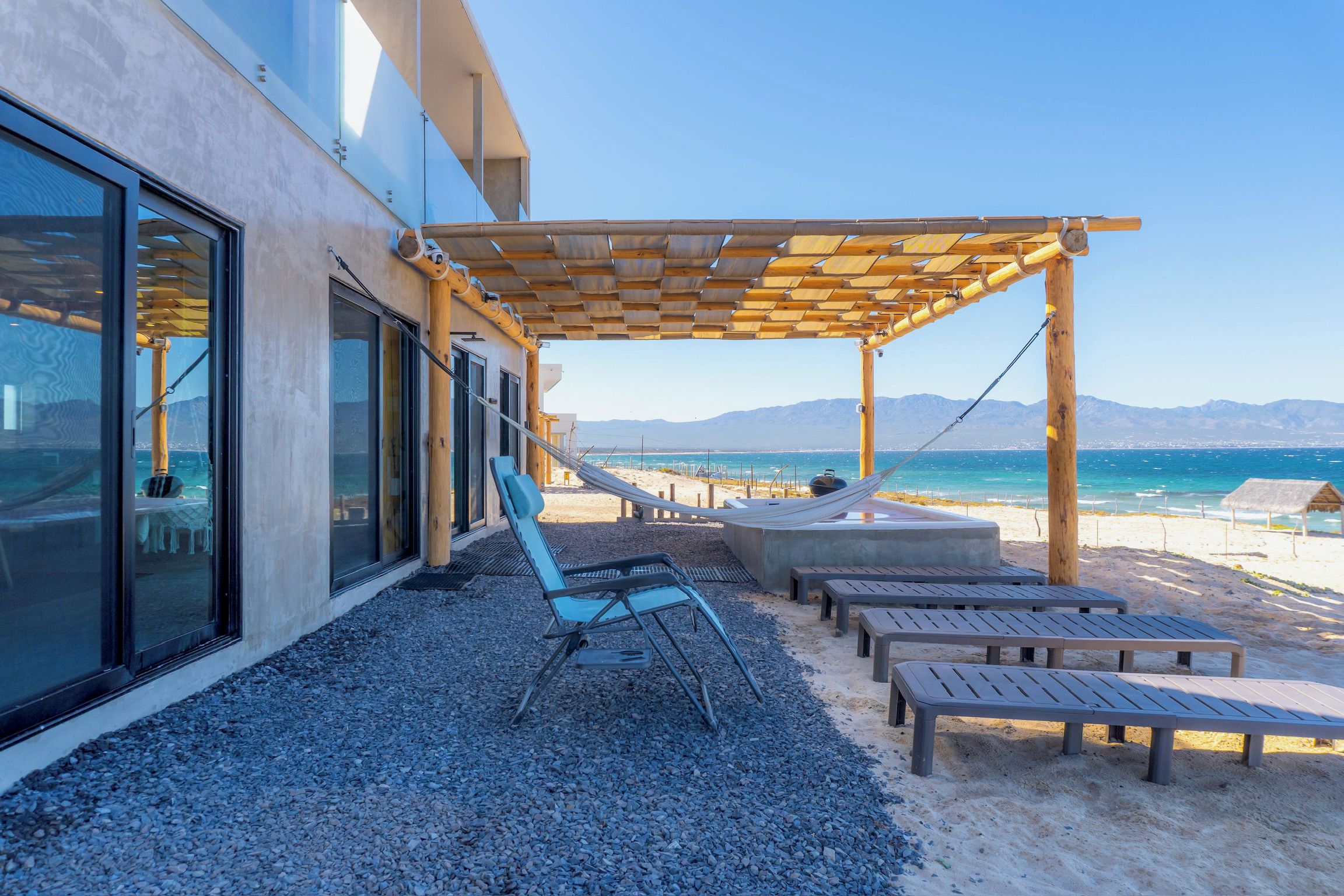 A beachside patio with a wooden pergola, lounge chairs, and a view of the ocean.