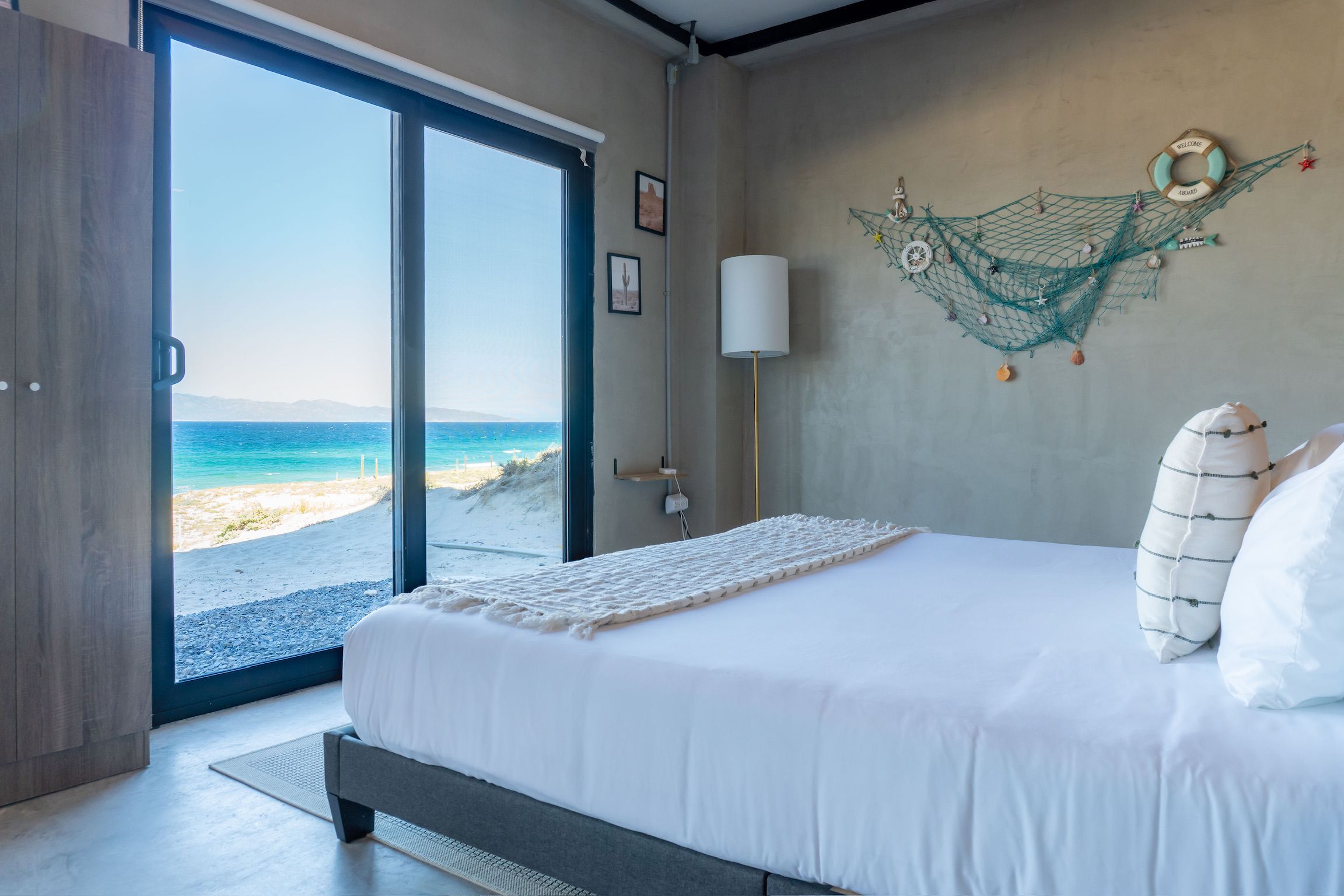 Un dormitorio con cama, mesita de noche, lámpara y vistas a la playa a través de una puerta corrediza.
