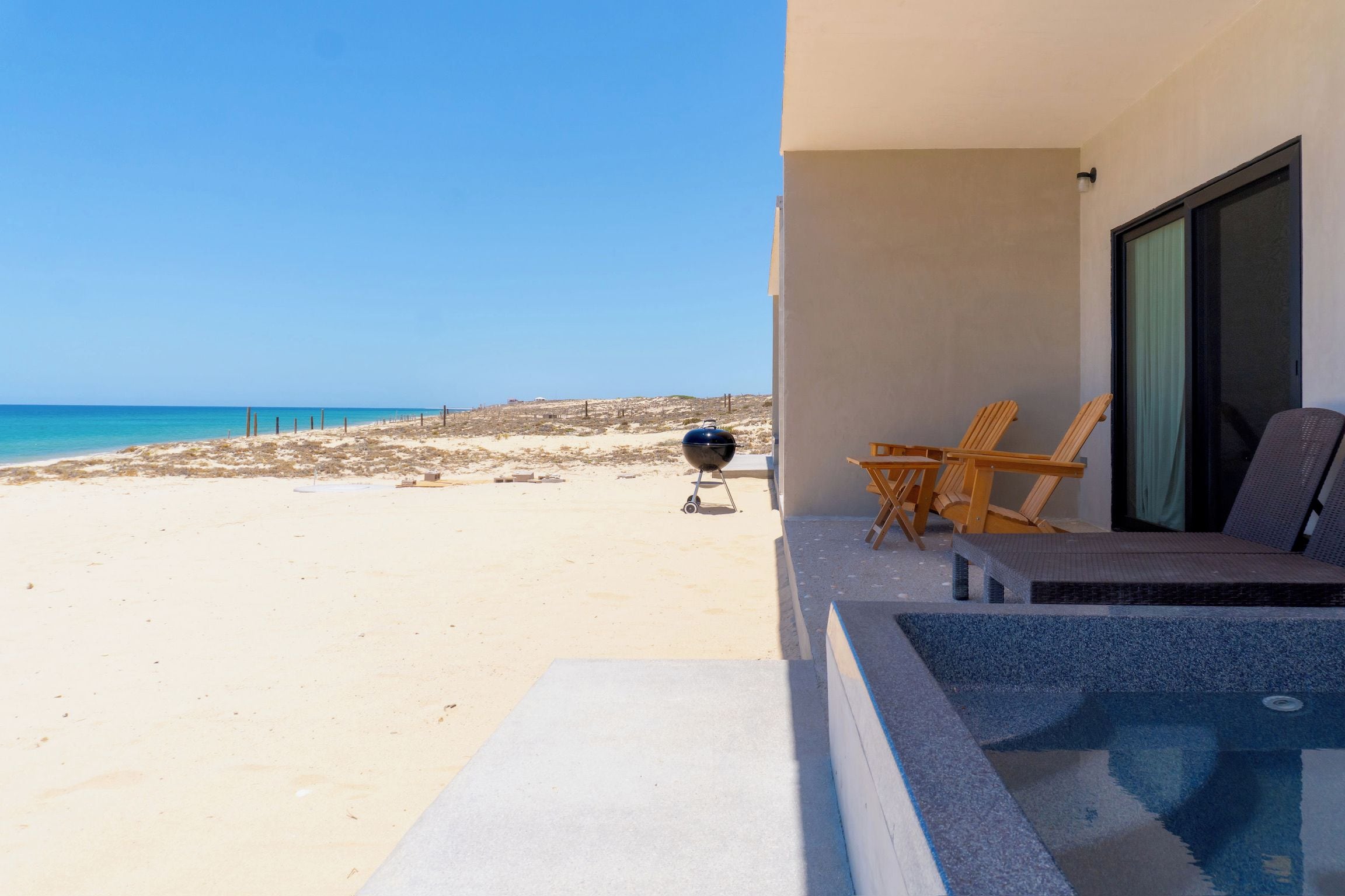 A beach view from a balcony with a barbecue grill, lounge chairs, and a pool area.